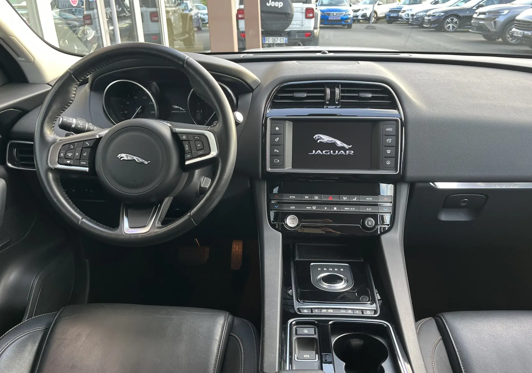Vue intérieure avant du Jaguar F-PACE blanc 2018, tableau de bord avec écran tactile et volant cuir noir multifonction.