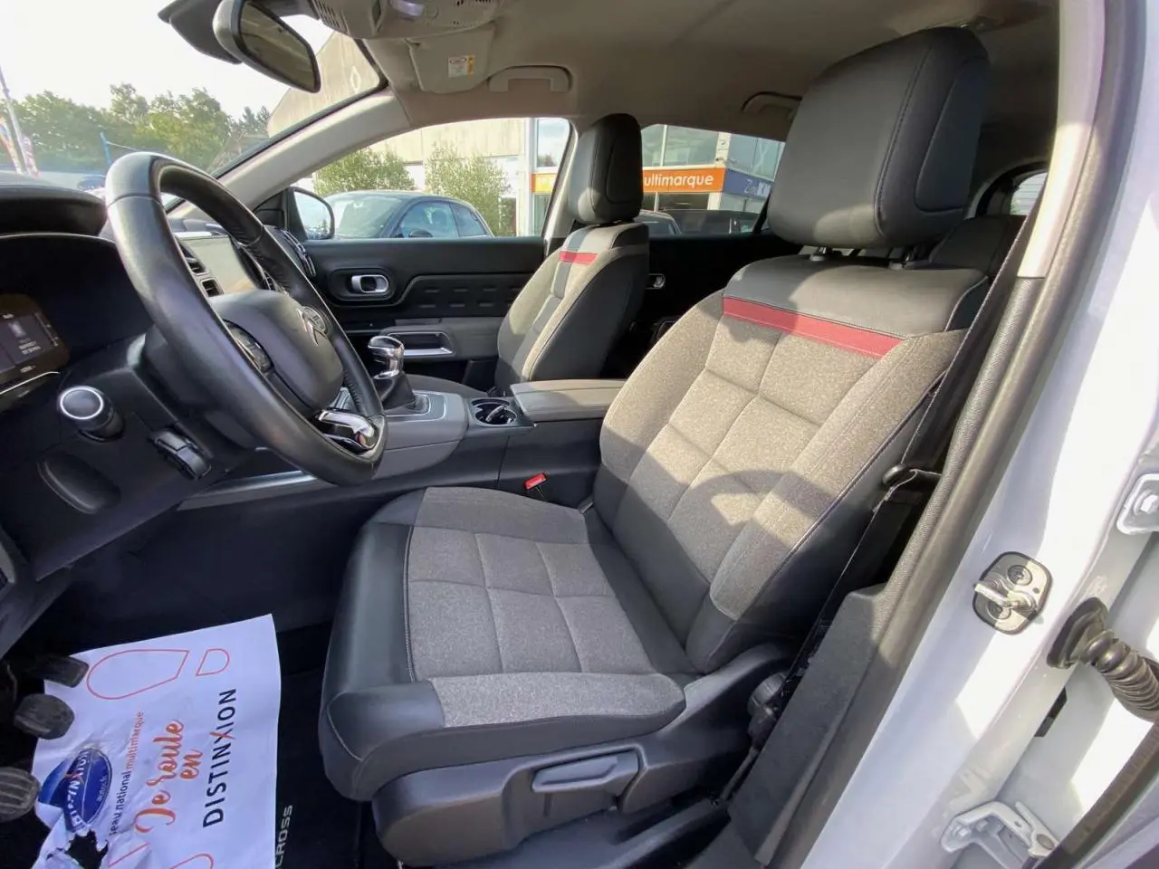 Intérieur avant droit du Citroën C5 Aircross blanc, sièges tissu et cuir gris avec bande rouge, volant et levier manuel visibles.