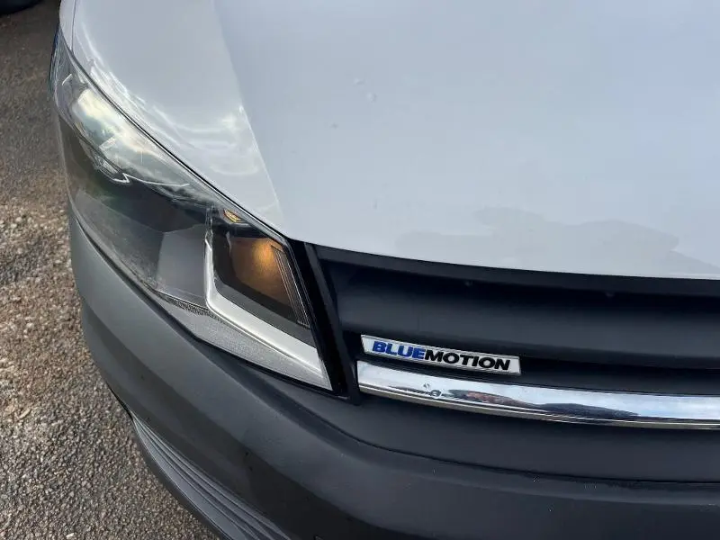 Gros plan sur le phare avant gauche et la calandre noire avec logo Bluemotion du Volkswagen Caddy Van blanc Candy.