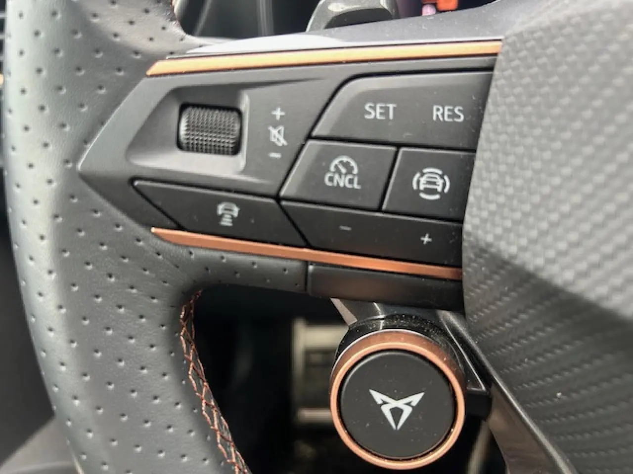 Gros plan sur le volant en cuir perforé gris avec commandes multifonctions et logo CUPRA cuivré au centre.