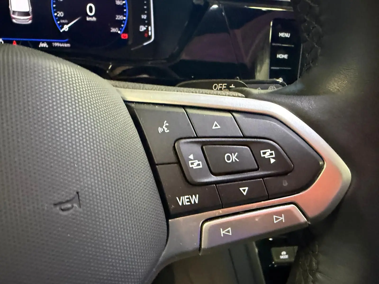 Gros plan sur les commandes multifonctions du volant cuir d’un Volkswagen Touran gris, tableau de bord numérique en arrière-plan.