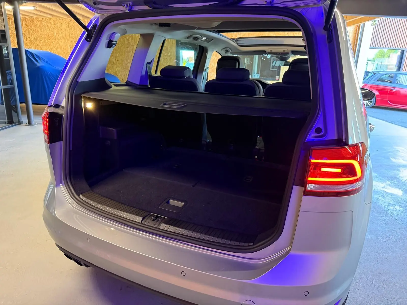 Vue arrière ouverte du coffre d'un Volkswagen Touran gris 2025 avec feux arrière LED allumés et toit ouvrant panoramique visible.