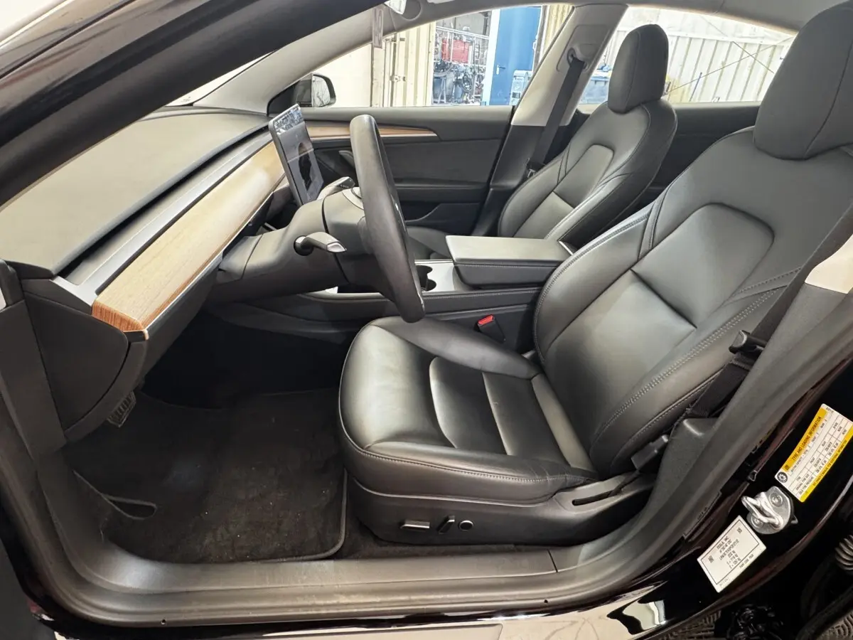 Intérieur côté conducteur de la Tesla Model 3 noire 2023, sièges cuir noir et tableau de bord bois clair.