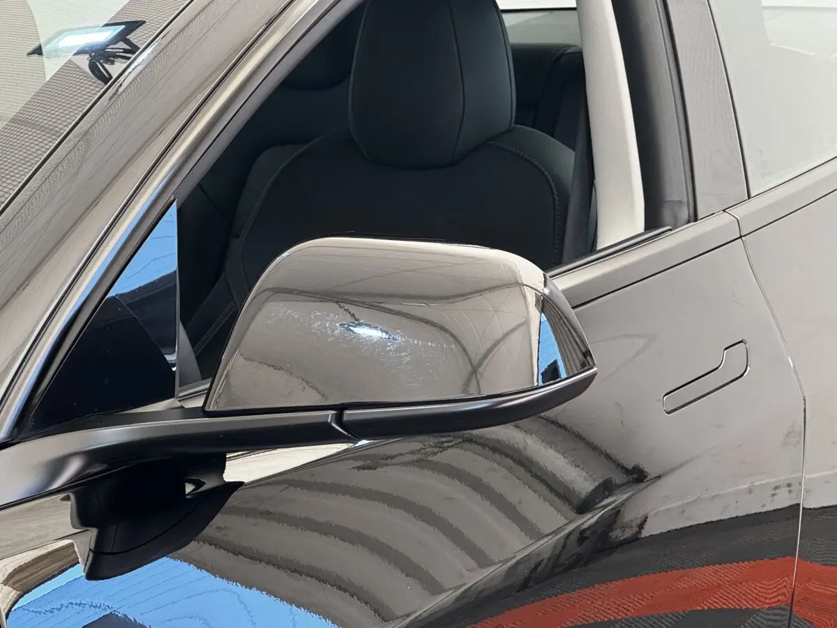 Gros plan sur le rétroviseur droit noir brillant d'une Tesla Model 3 Grande Autonomie 2023, avec porte et siège visibles.