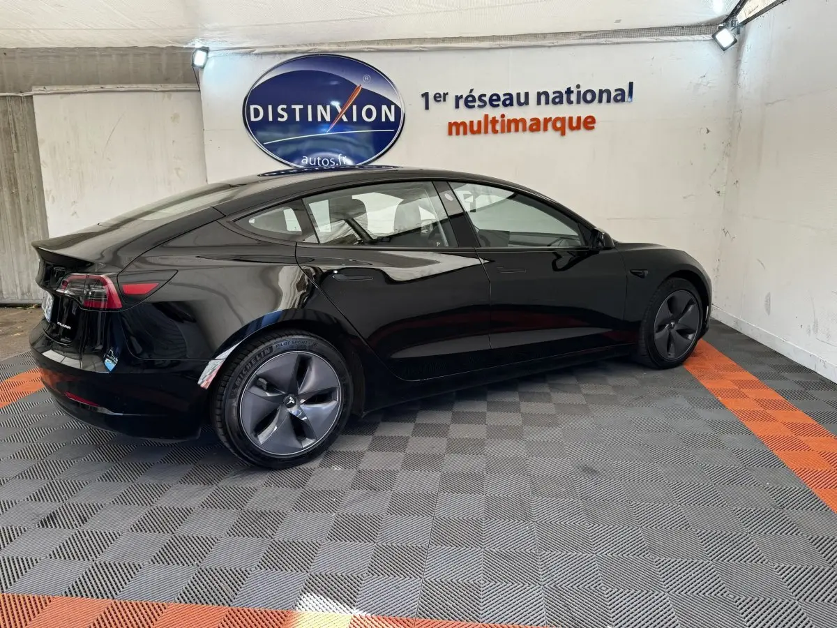 Tesla Model 3 Grande Autonomie noir, vue 3/4 arrière côté gauche dans un showroom avec sol gris et orange.