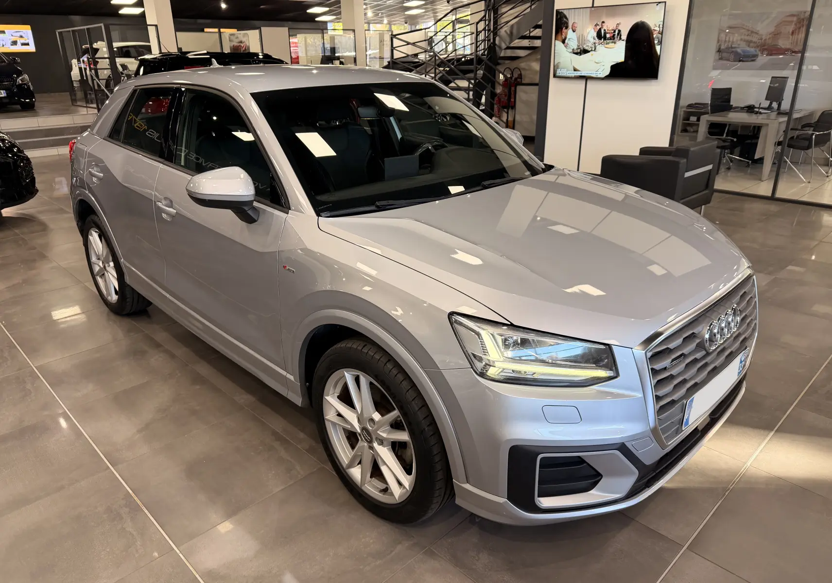 Audi Q2 gris en 3/4 avant droit dans un showroom, avec phares LED et jantes alliage visibles.