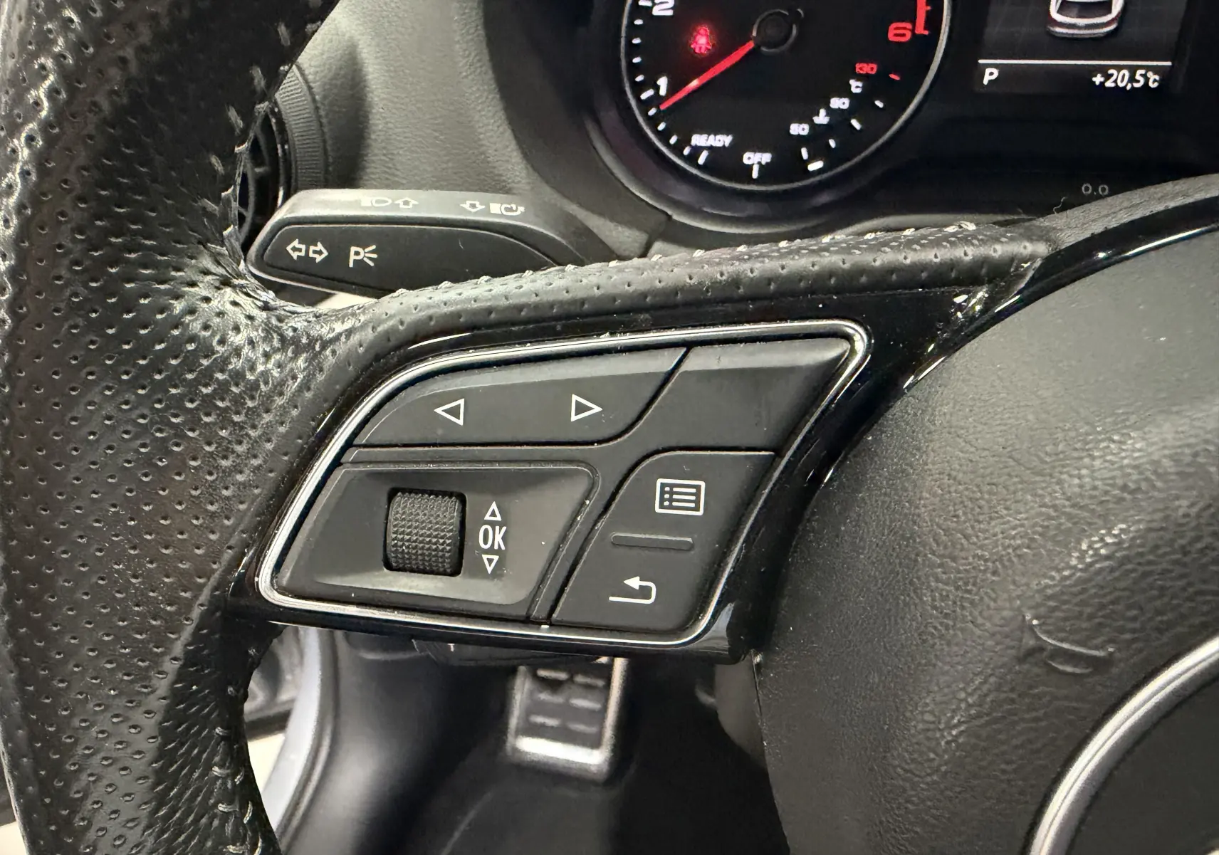Gros plan sur les commandes multifonctions à gauche du volant perforé noir de l'Audi Q2 gris, avec compte-tours en arrière-plan.