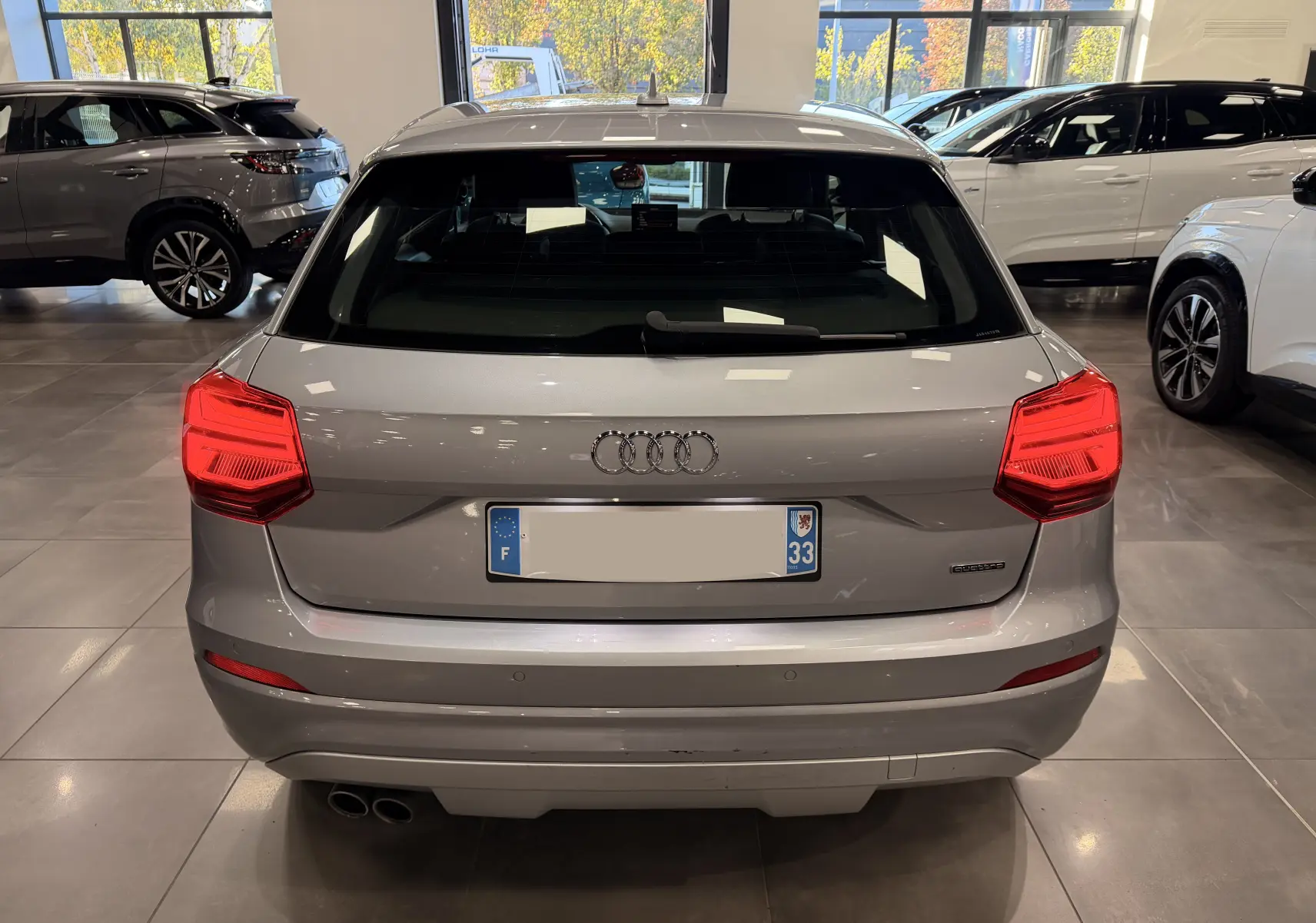 Vue arrière d'un Audi Q2 gris 2017 en intérieur, avec feux arrière allumés et double sortie d'échappement visible.