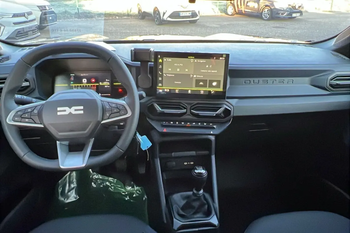 Vue intérieure du tableau de bord du Dacia Duster 2025 avec volant multifonction, écran tactile et levier de vitesse manuel.