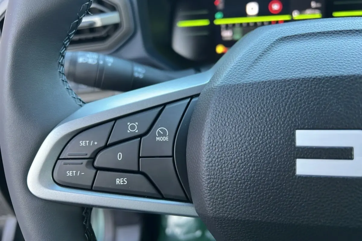 Gros plan sur les commandes au volant côté gauche du Dacia Duster 2025 vert cèdre, avec affichage numérique en arrière-plan.