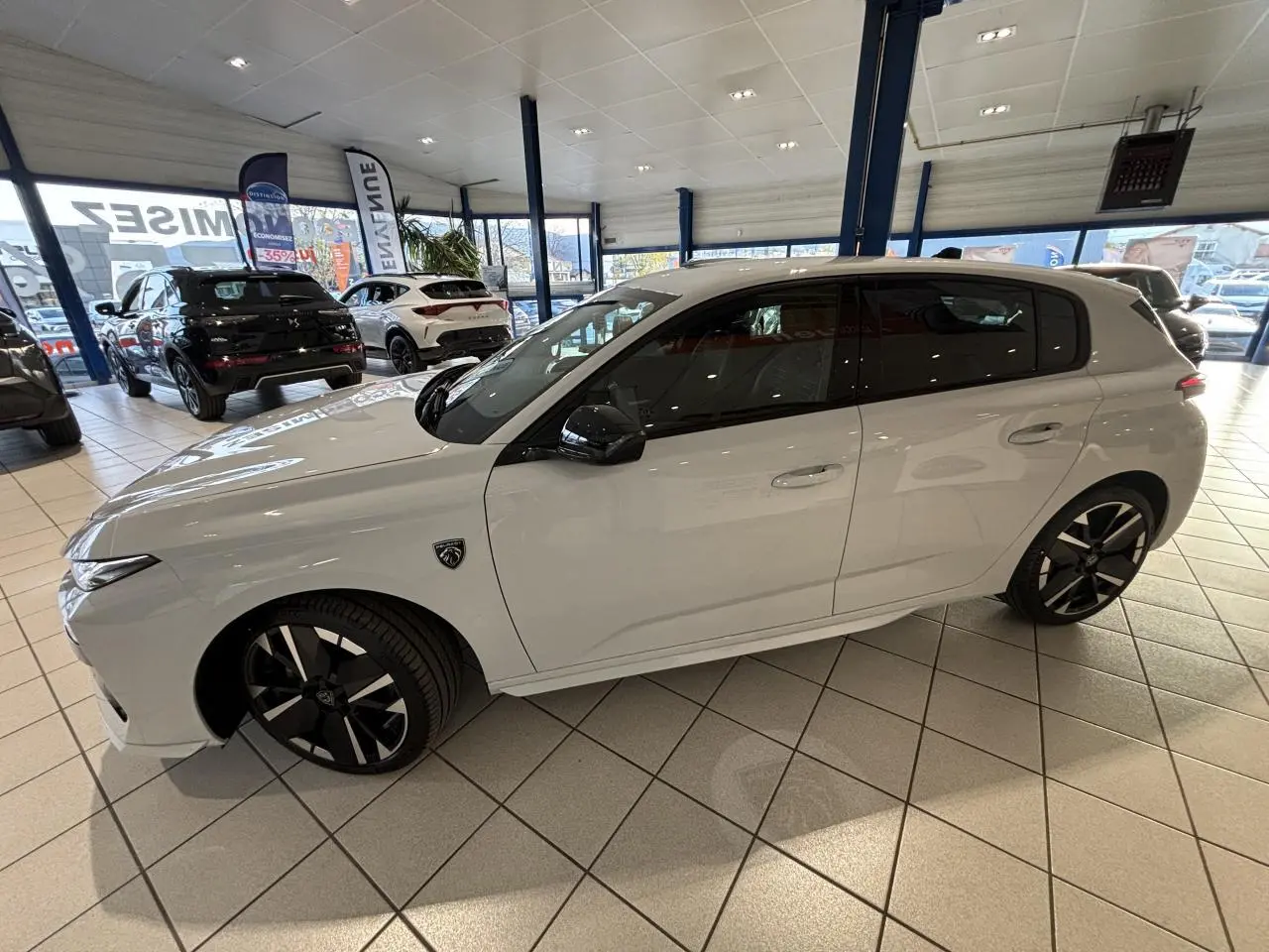 Peugeot 308 blanche vue de profil côté gauche en intérieur, avec jantes alliage noires et blason distinctif sur aile avant.