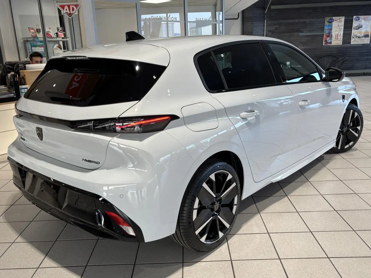 Vue 3/4 arrière droite d'une Peugeot 308 Hybrid blanche avec jantes alliage bicolores et vitres teintées.