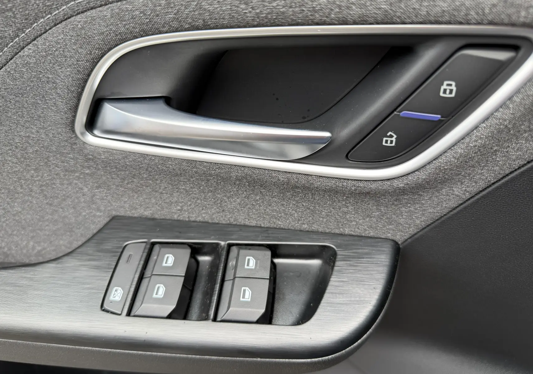 Poignée de porte intérieure et commandes des vitres en tissu gris et plastique noir du MG ZS 1.5L Hybrid+ 2026 côté conducteur.