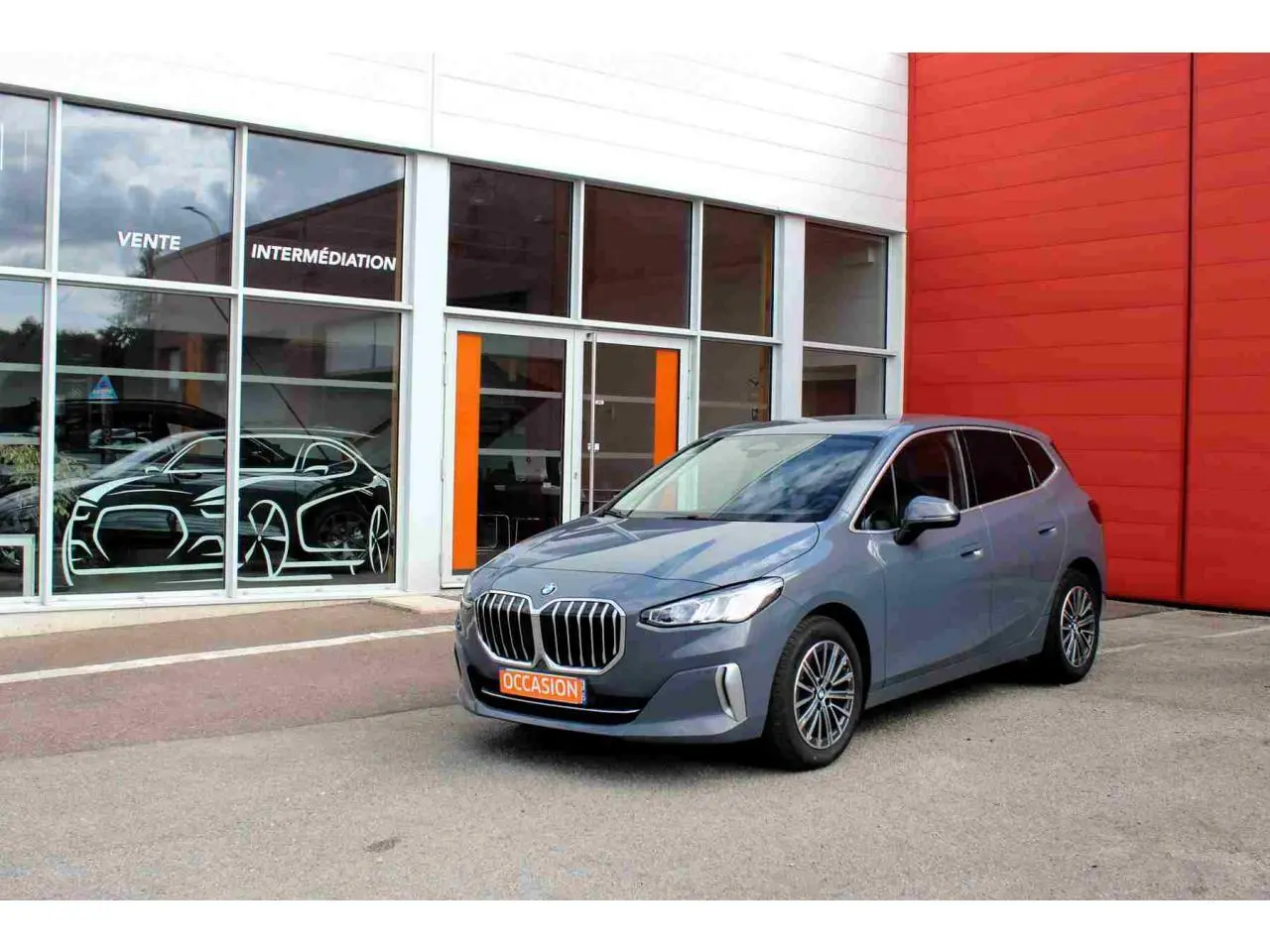 BMW Série 2 Active Tourer gris vue 3/4 avant avec calandre chromée, stationnée devant un bâtiment orange et blanc.