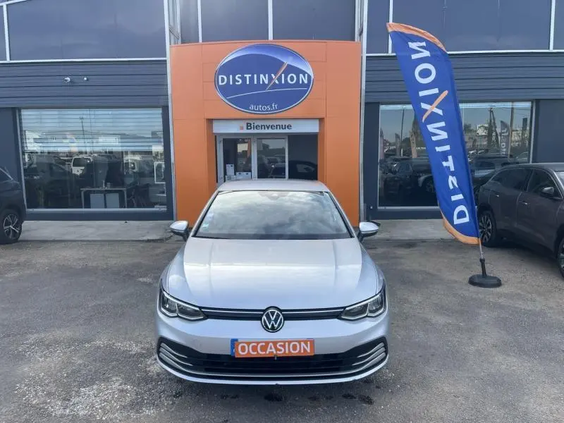 Vue avant d'une Volkswagen Golf 1.5 eTSI argent métallisé, devant un showroom avec drapeau Distinxion.