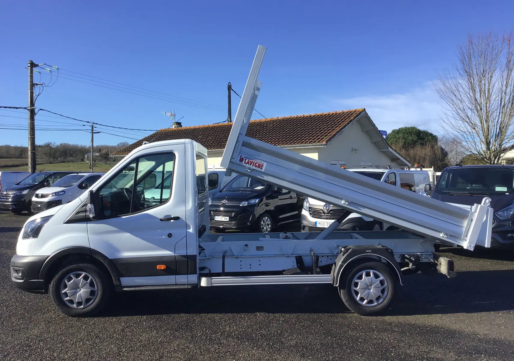 Vue de profil côté gauche d'un Ford Transit blanc avec benne basculante levée sur un parking extérieur.