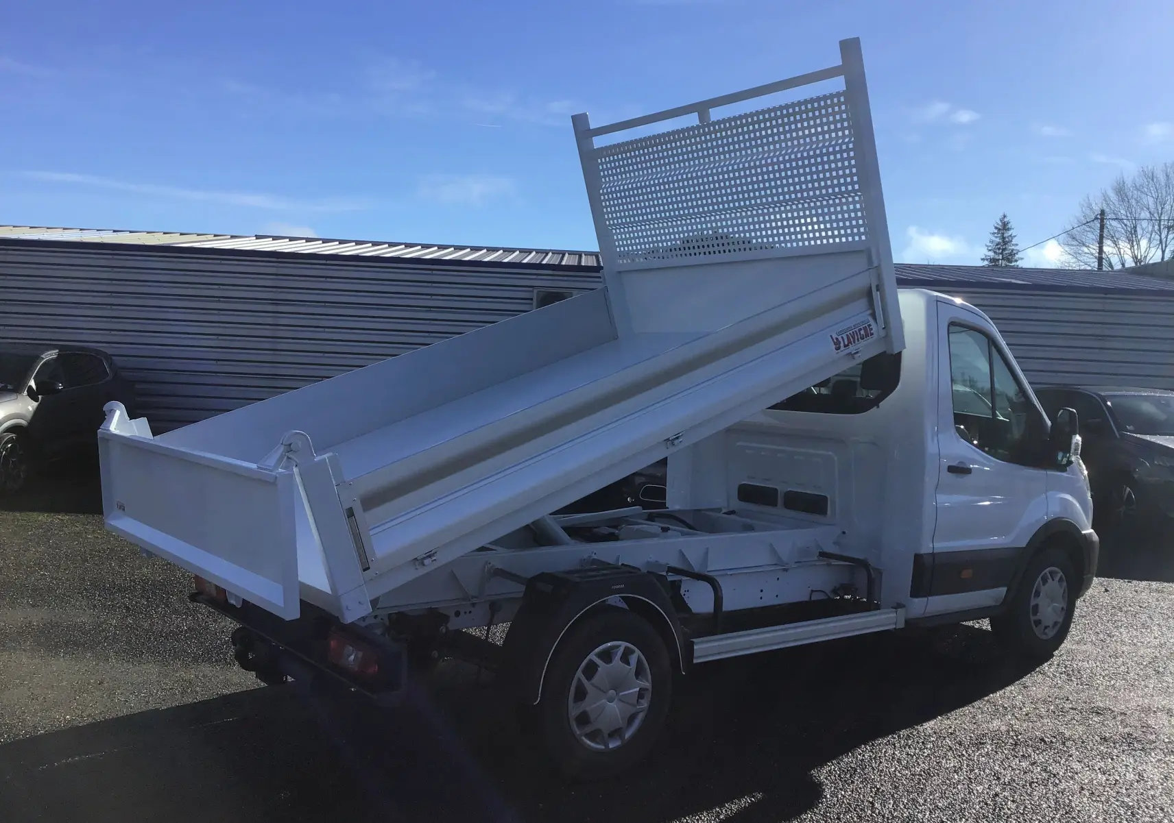 Vue 3/4 arrière droite du Ford Transit Châssis cabine blanc avec benne basculante levée sous un ciel bleu.