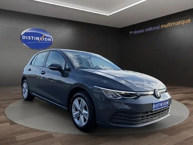 Volkswagen Golf gris clair vue 3/4 avant droit en showroom avec logo Distinxion en arrière-plan.