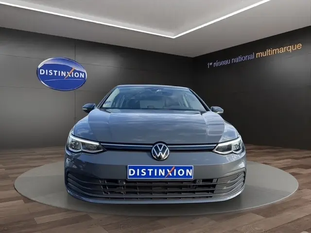 Vue frontale d'une Volkswagen Golf gris clair 2023 exposée en showroom avec logo Distinxion en arrière-plan.