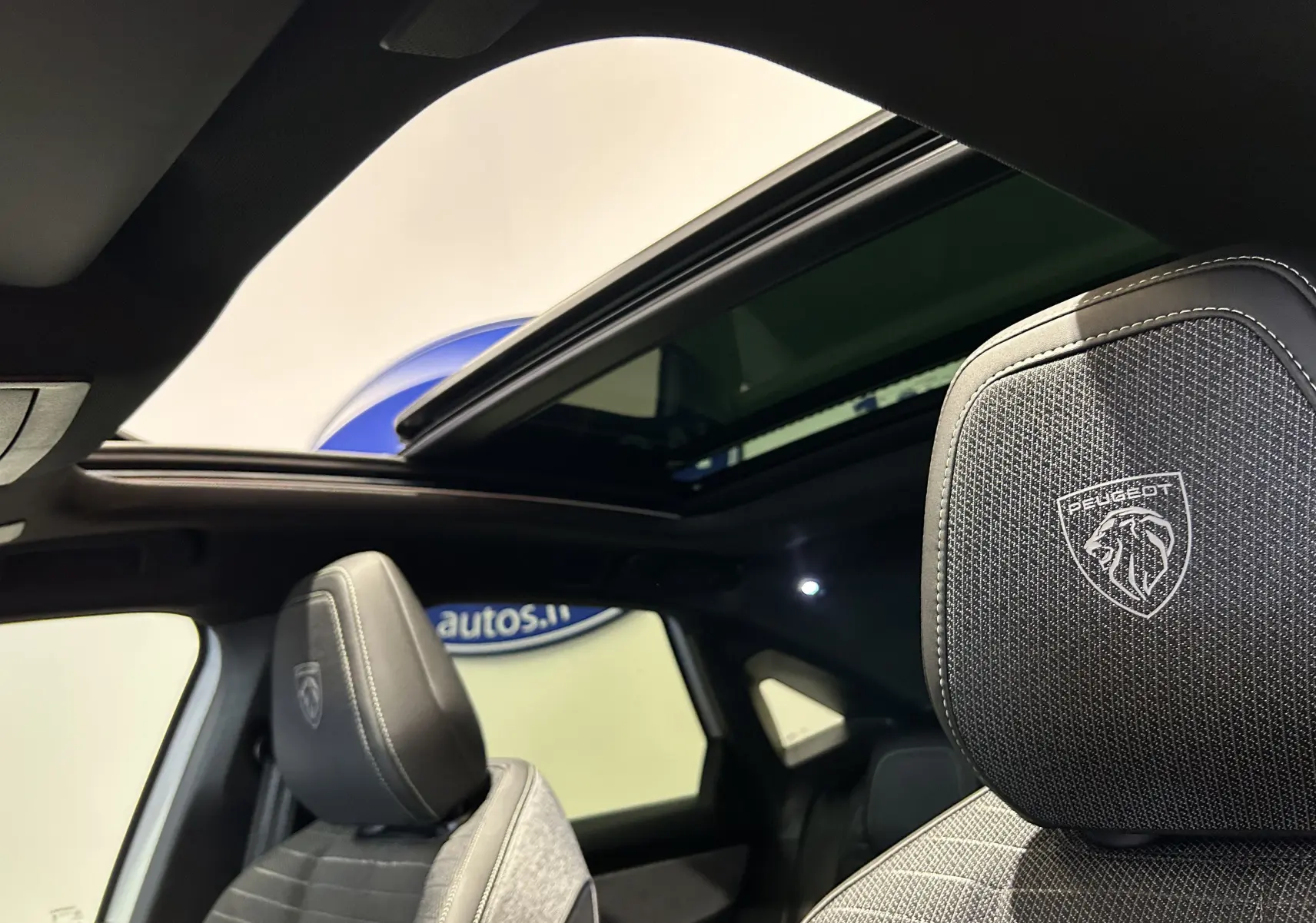Intérieur du Peugeot 3008 Hybrid 2026, vue sur les sièges avant gris avec logo Peugeot et toit ouvrant panoramique ouvert.