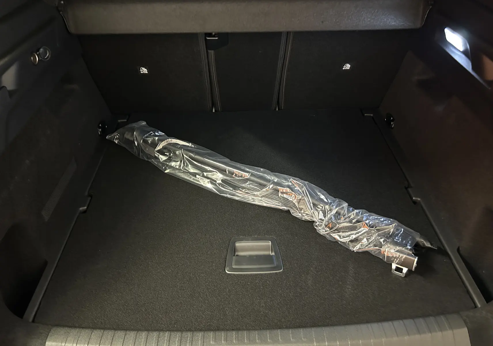 Coffre noir du Peugeot 3008 Hybrid 2026, vue plongeante, avec un accessoire emballé en plastique transparent.