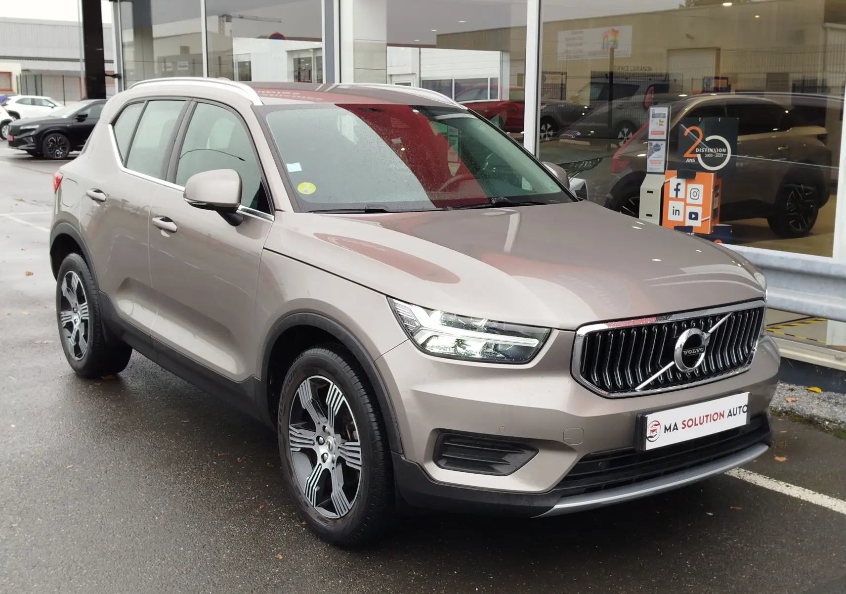 Vue 3/4 avant droit d'un Volvo XC40 beige 2020 avec calandre verticale et jantes bi-ton, stationné devant un showroom.