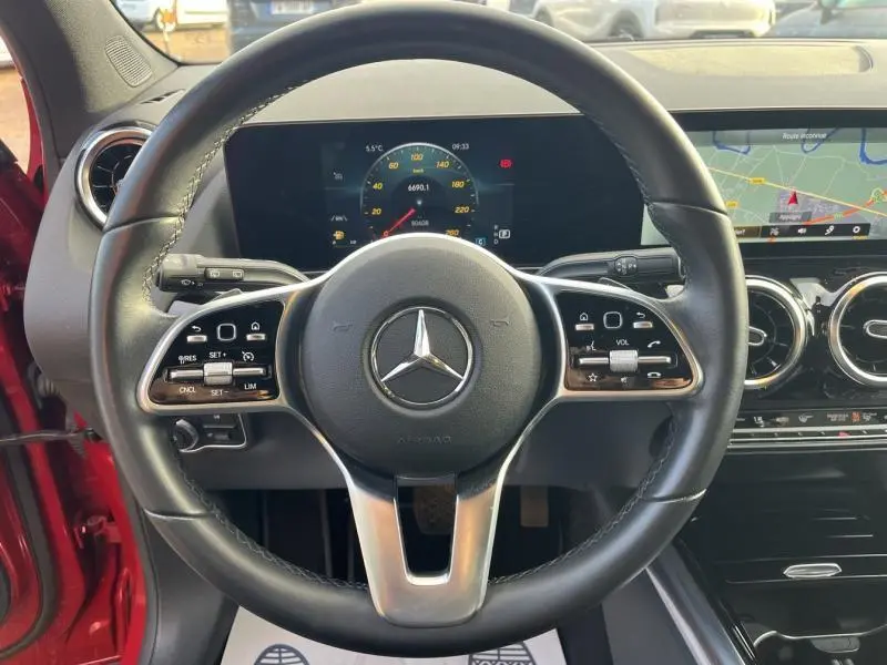 Vue intérieure centrée sur le volant cuir multifonction d'une Mercedes GLA rouge Patagonie, avec tableau de bord digital et écran tactile GPS visible.