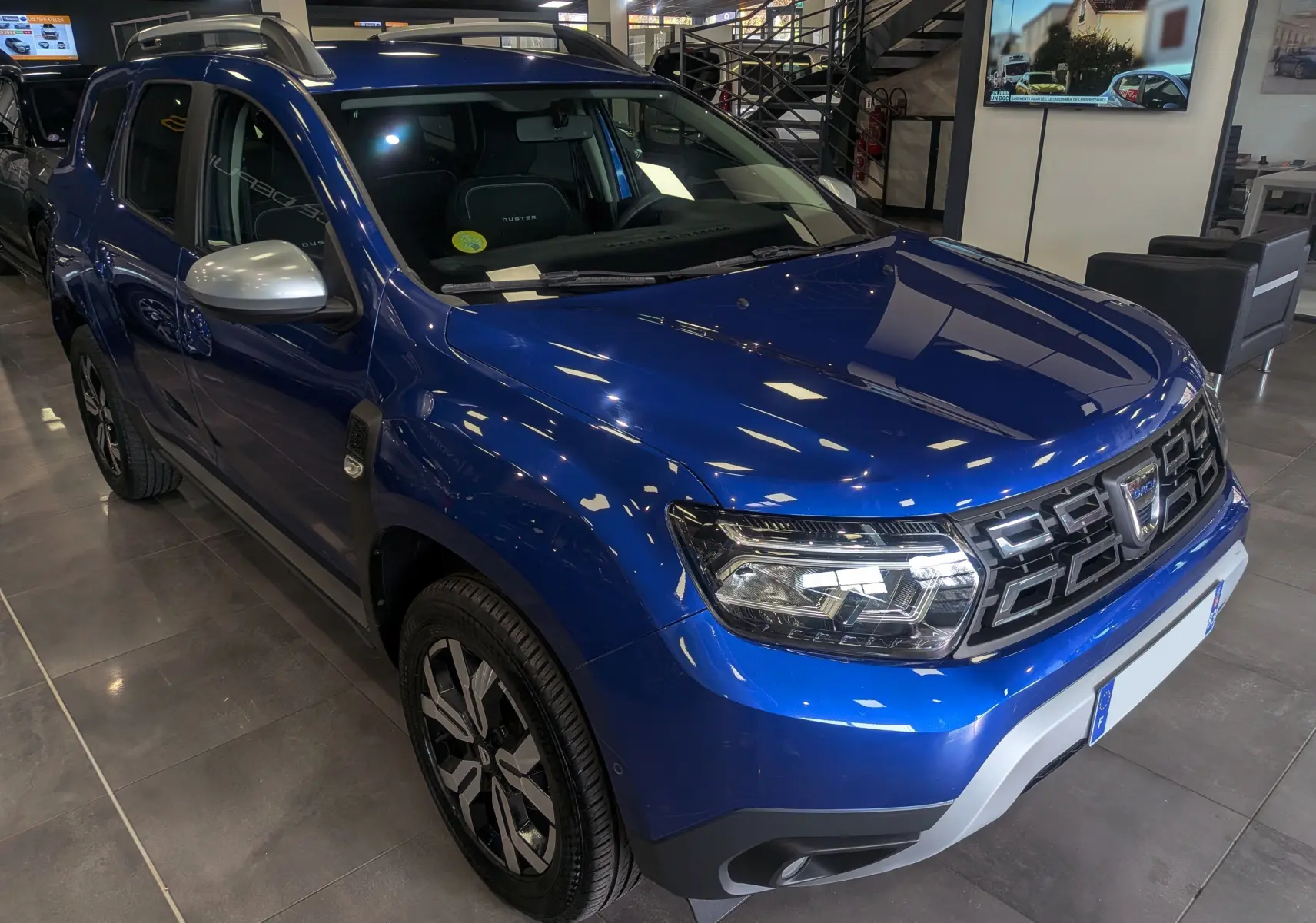 Vue 3/4 avant gauche du Dacia Duster 2022 bleu Iron avec jantes bi-ton et rétroviseurs argentés en showroom.