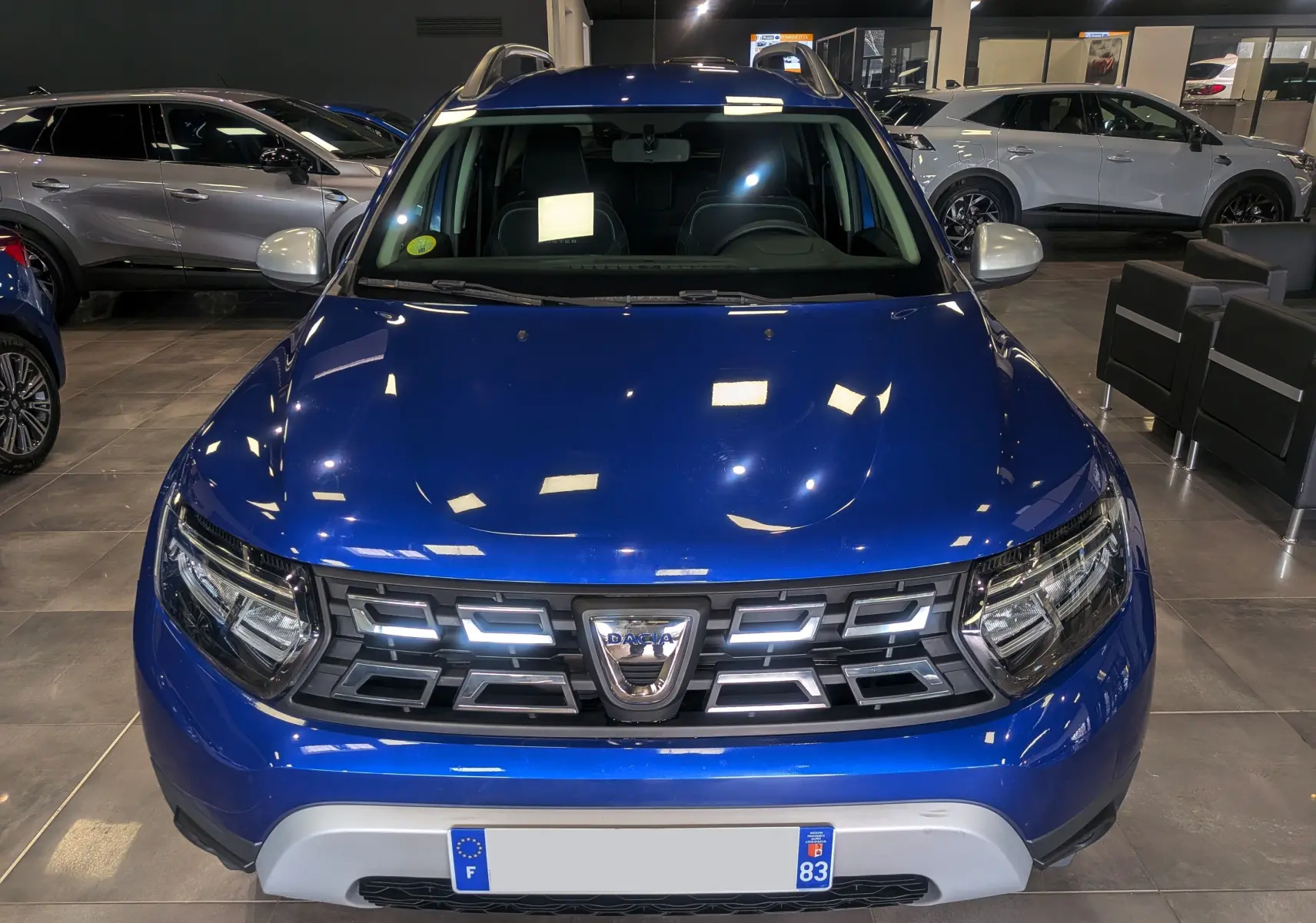 Vue avant d'un Dacia Duster 2022 Bleu Iron en showroom, avec calandre distinctive et phares LED allumés.
