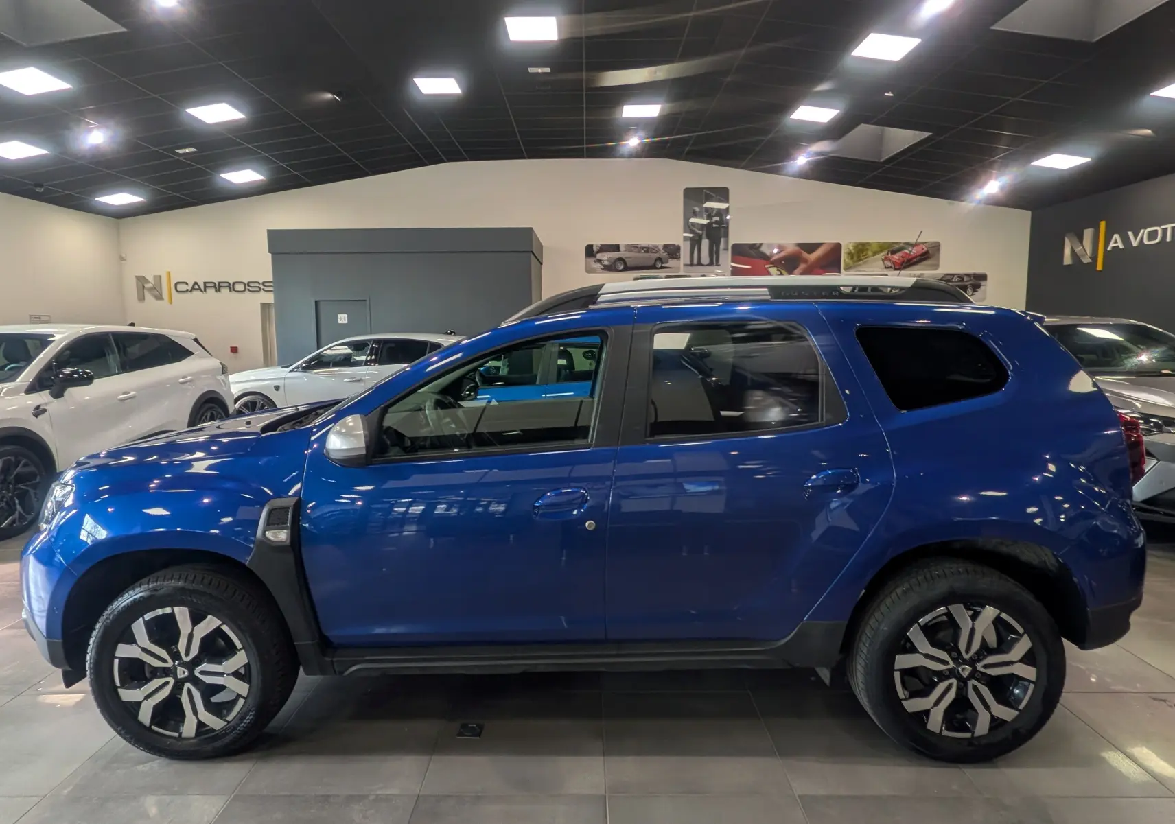 Vue de profil côté gauche d'un Dacia Duster 2022 Bleu Iron avec jantes bi-ton et barres de toit noires.
