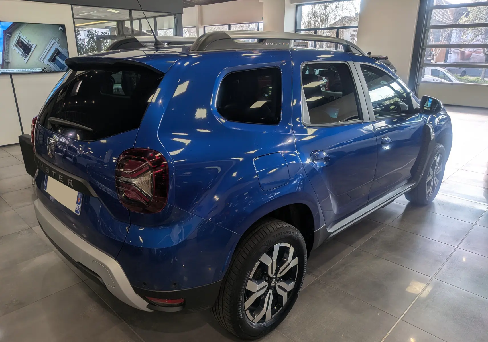 Vue 3/4 arrière droite du Dacia Duster 2022 Bleu Iron avec barres de toit et jantes bi-ton en showroom.