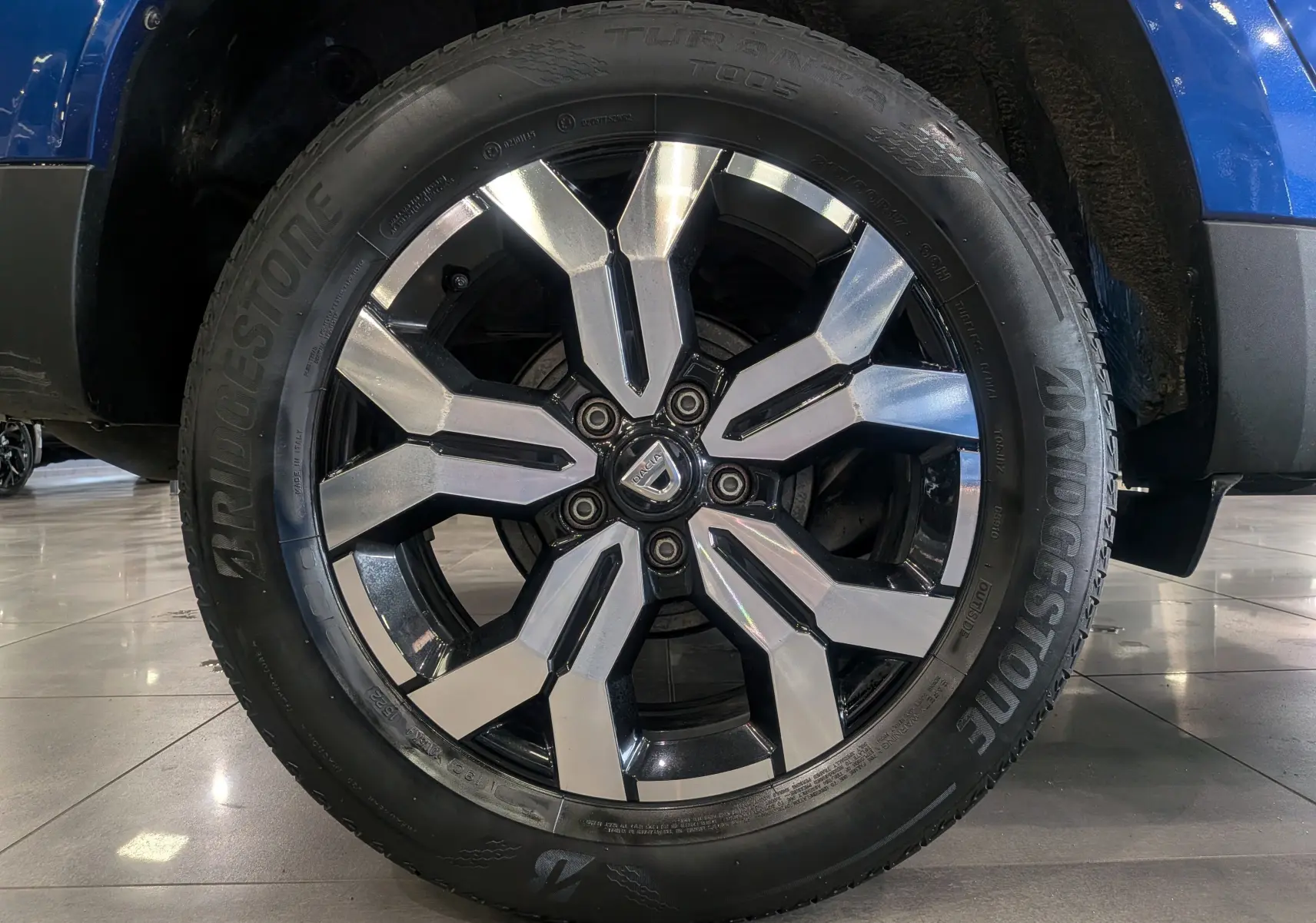Gros plan sur la jante alliage bicolore du Dacia Duster Bleu Iron, mettant en valeur le logo central et le pneu Bridgestone.