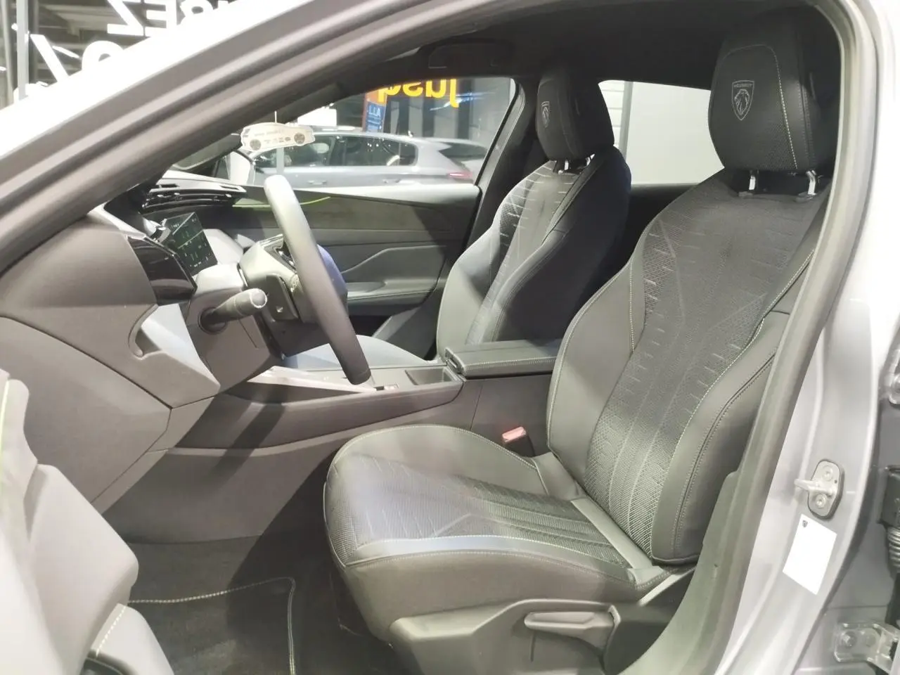 Vue intérieure côté conducteur de la Peugeot 308 gris Artense, sièges noirs avec surpiqûres vert Adamite et logo Peugeot brodé.