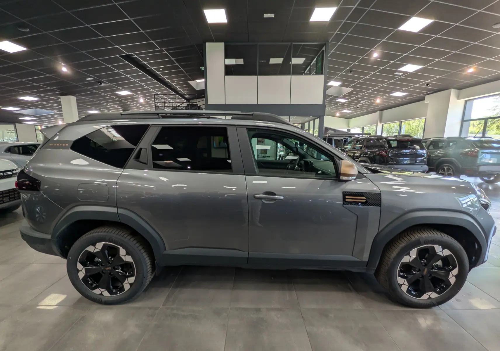 Vue de profil côté droit du Dacia Bigster Hybrid-G 150 4x4 gris schiste avec jantes biton noires en showroom.