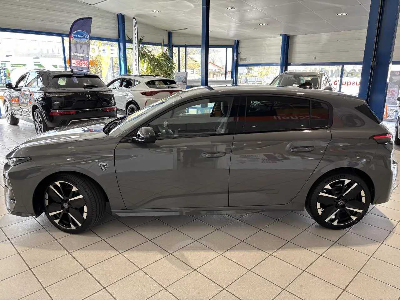Vue de profil côté gauche d’une Peugeot 308 gris selenium en showroom avec jantes alliage noires et blason sur aile avant.