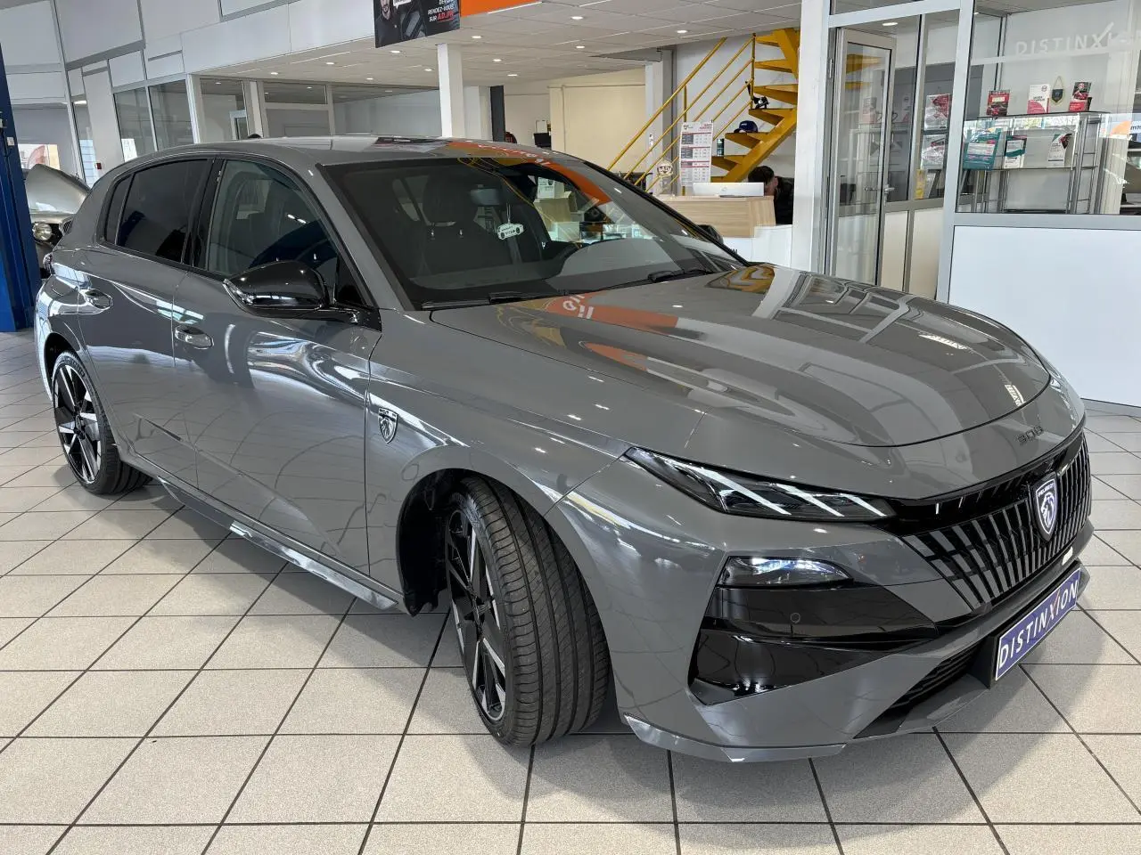 Peugeot 308 gris selenium vue 3/4 avant droit en showroom avec calandre noire et jantes alliage bi-ton.
