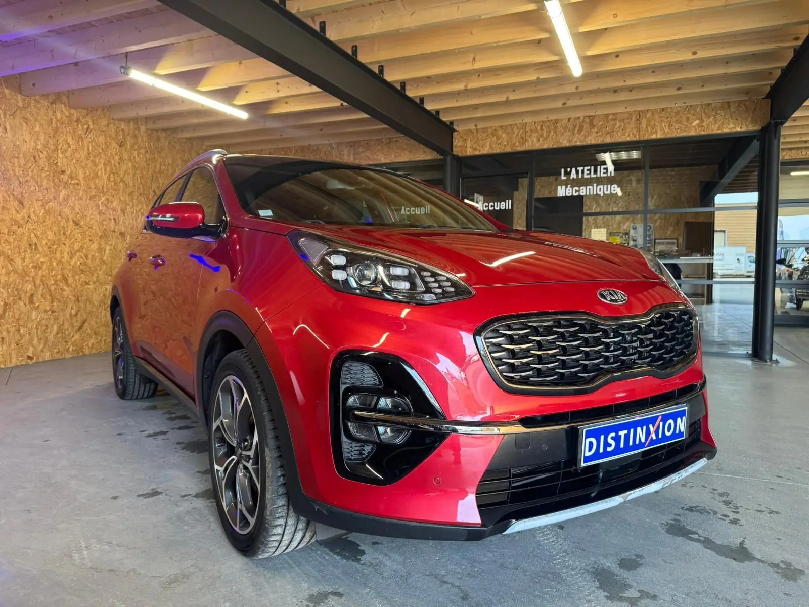 KIA Sportage rouge 2019 en 3/4 avant droit, avec calandre noire et jantes alliage visibles en intérieur d'atelier.