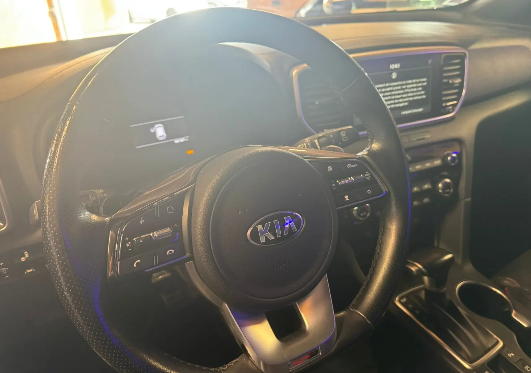 Vue intérieure centrée sur le volant noir perforé du Kia Sportage 1.6 CRDI, avec tableau de bord et écran tactile en arrière-plan.