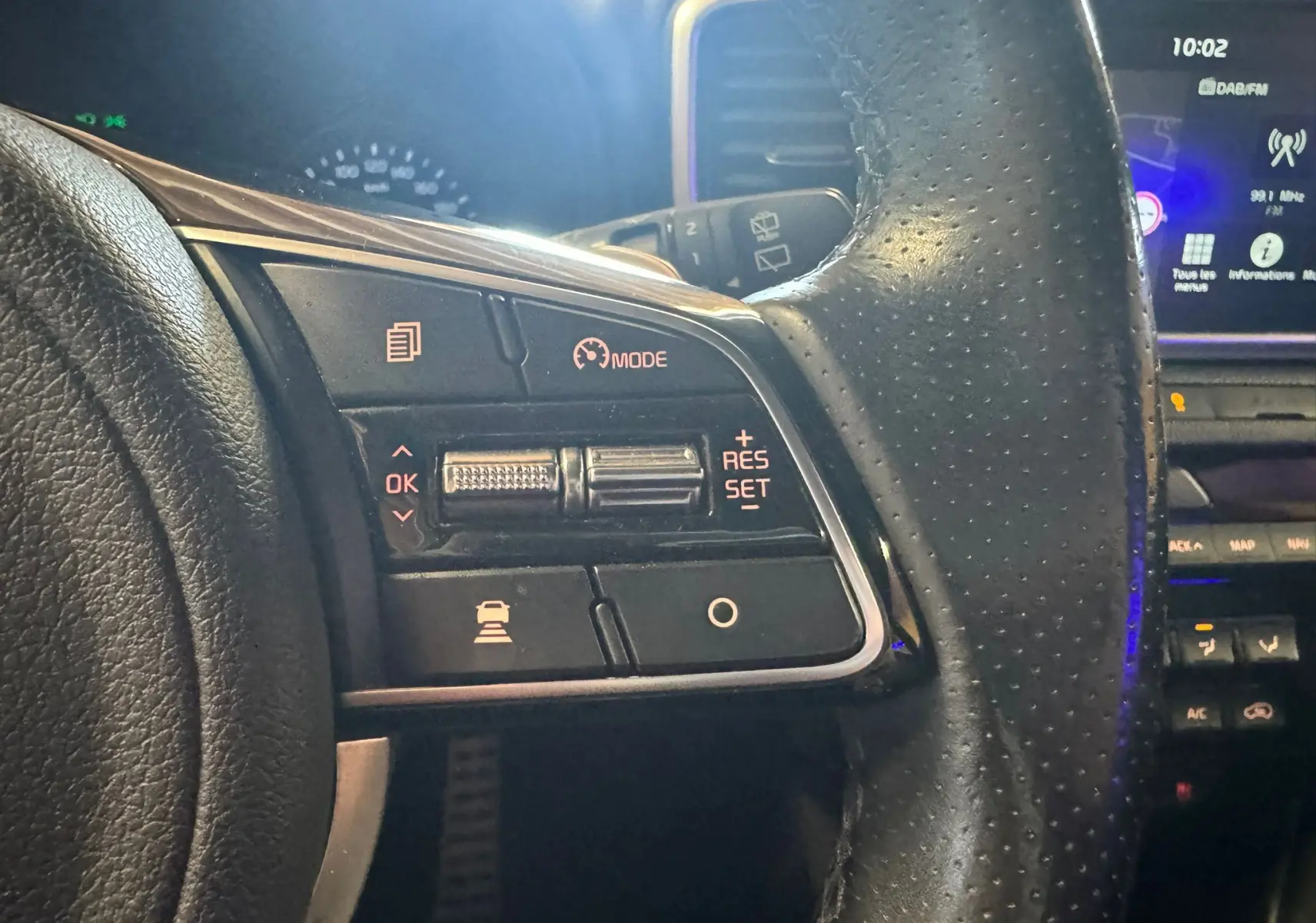 Gros plan sur les commandes au volant en cuir noir du Kia Sportage 1.6 CRDI 136 GT Line, avec affichage digital en arrière-plan.