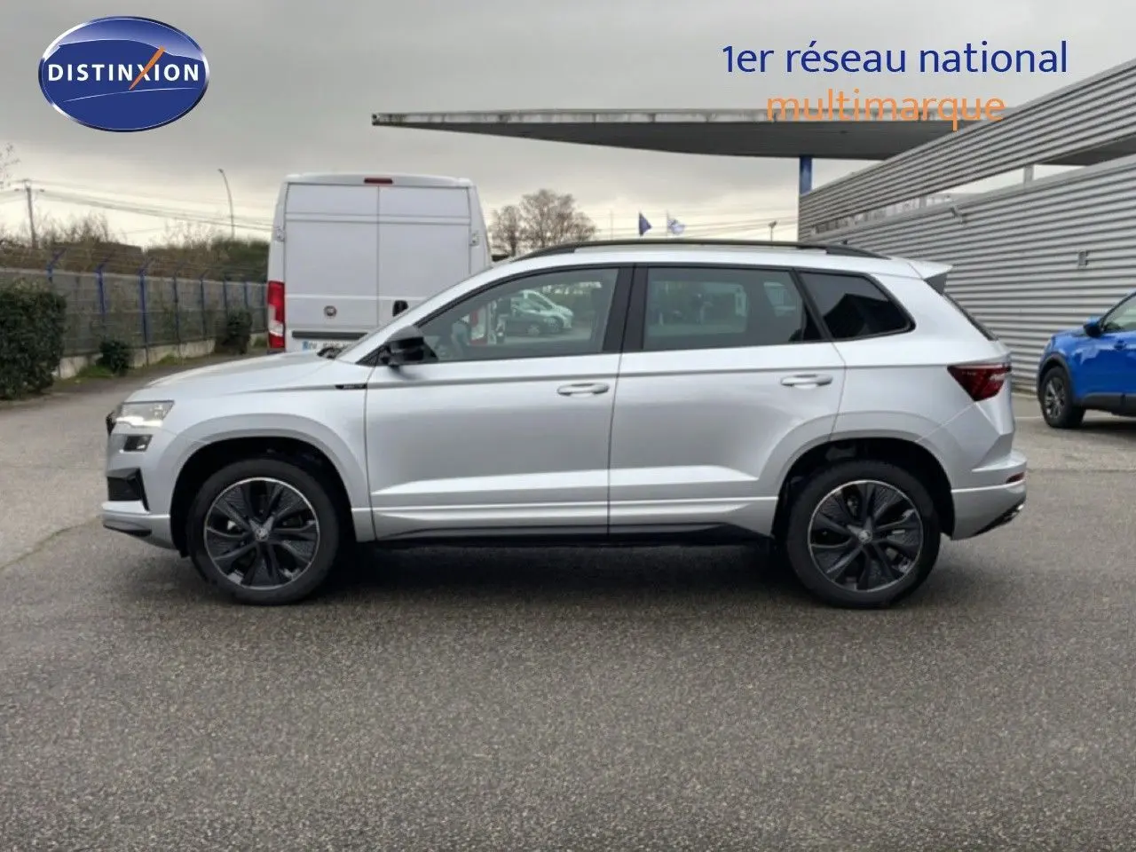Vue de profil côté gauche d'un Skoda Karoq gris argent métallisé avec jantes foncées sur parking extérieur.