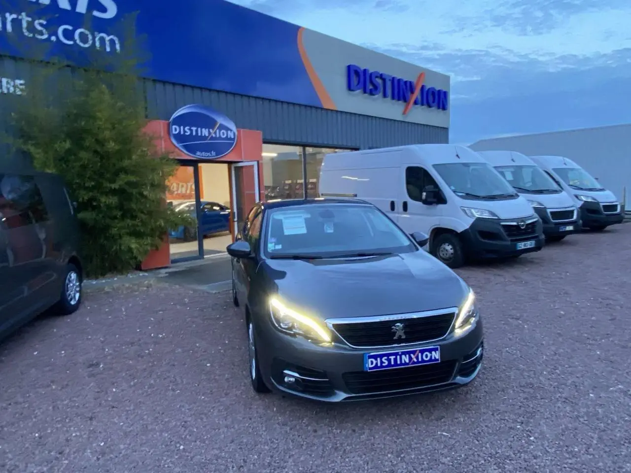 Peugeot 308 gris clair vue 3/4 avant avec phares allumés, stationnée devant un concessionnaire Distinxion.