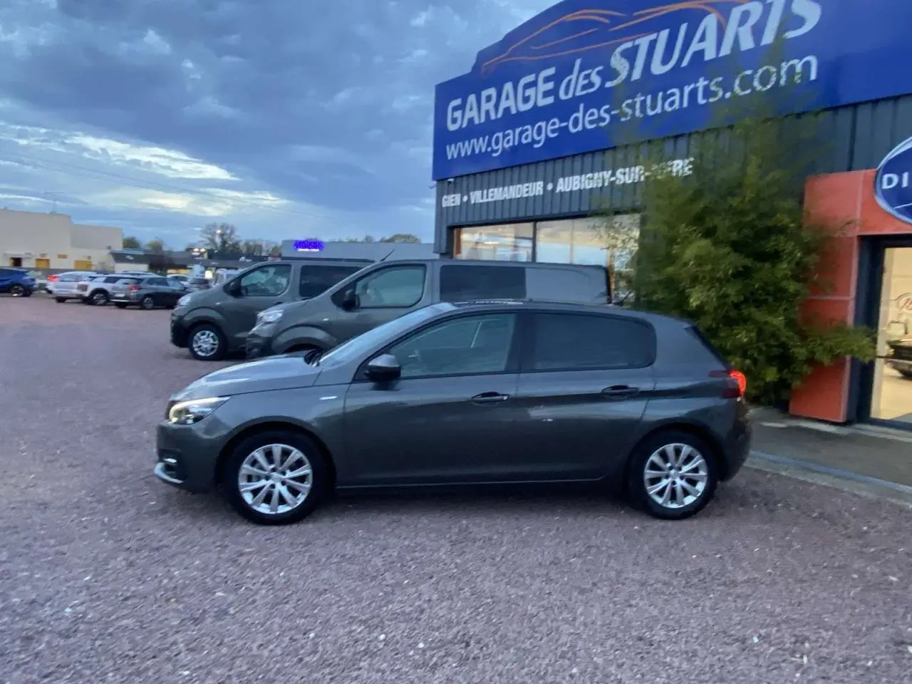 Peugeot 308 gris clair vue de profil côté gauche, avec jantes aluminium 16 pouces, stationnée devant un garage.