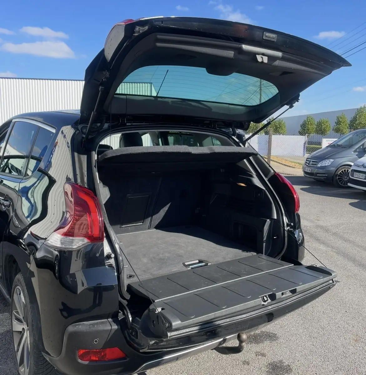 Peugeot 3008 noir vue 3/4 arrière gauche avec coffre ouvert et hayon à double ouverture sous un ciel bleu.