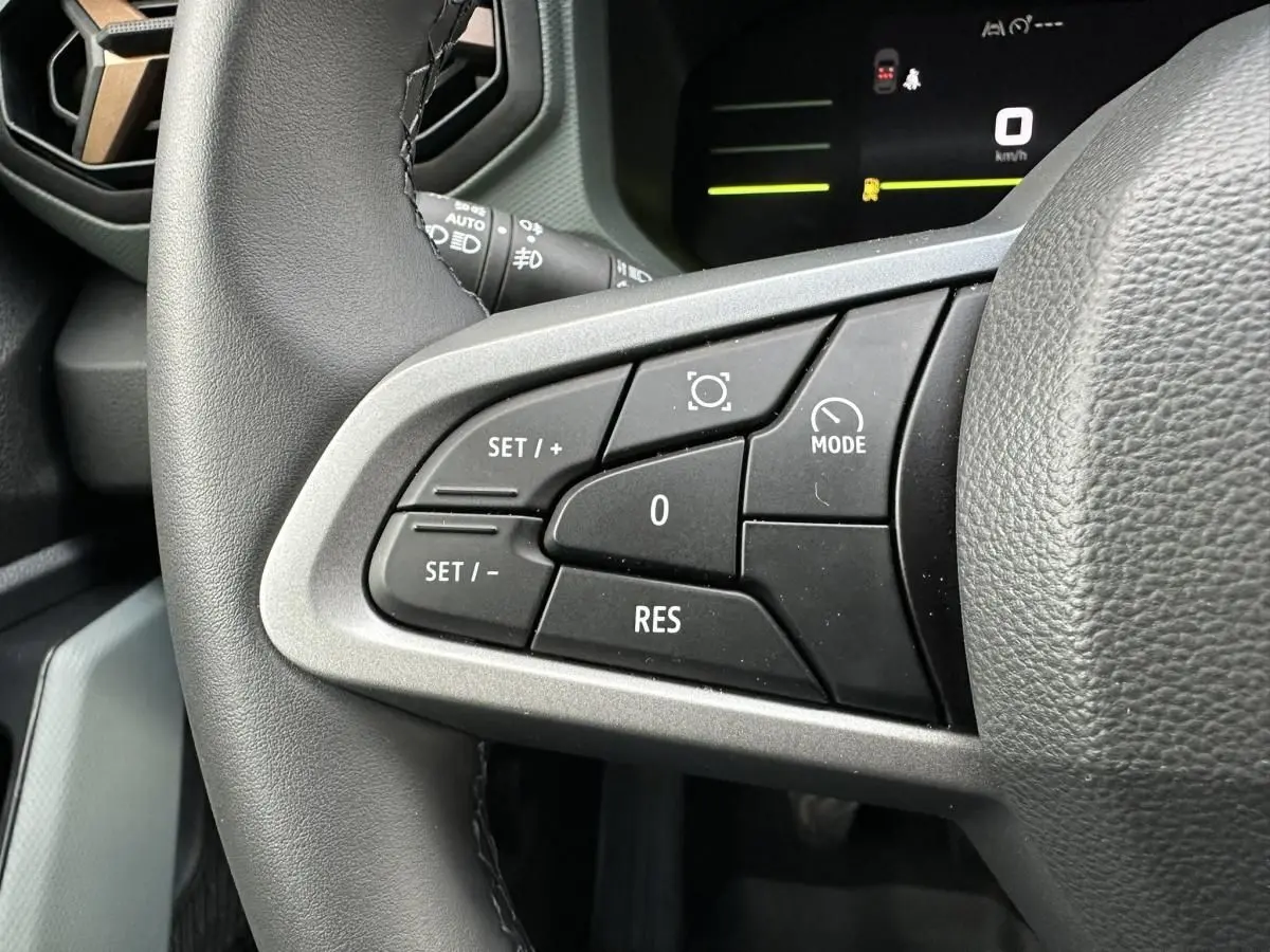 Gros plan sur les commandes au volant à gauche du Dacia Duster blanc Glacier 2025, avec affichage numérique en arrière-plan.