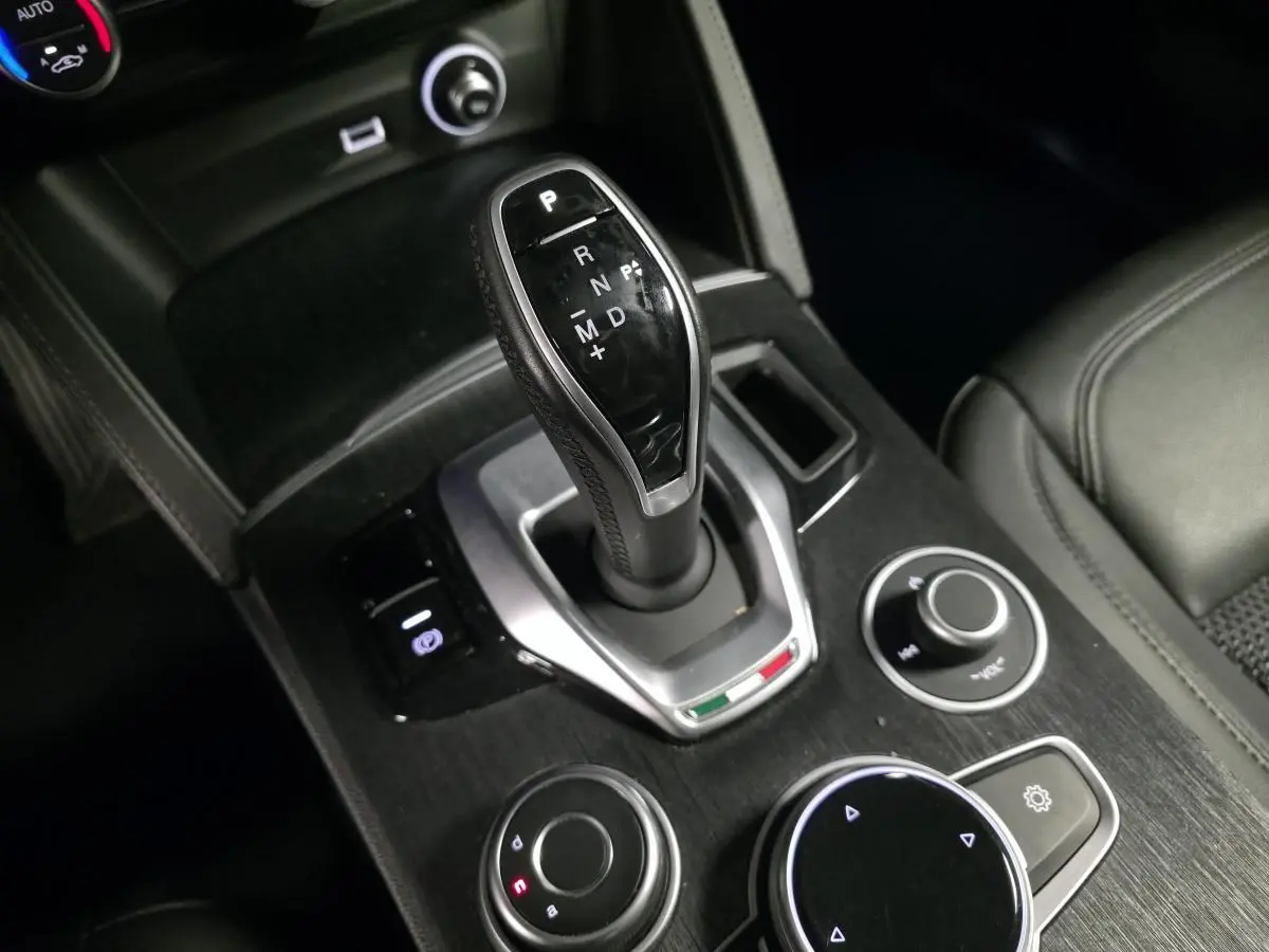 Gros plan sur la console centrale noire de l'Alfa Romeo Stelvio 2.2 D avec levier de vitesse automatique et commandes rotatives.