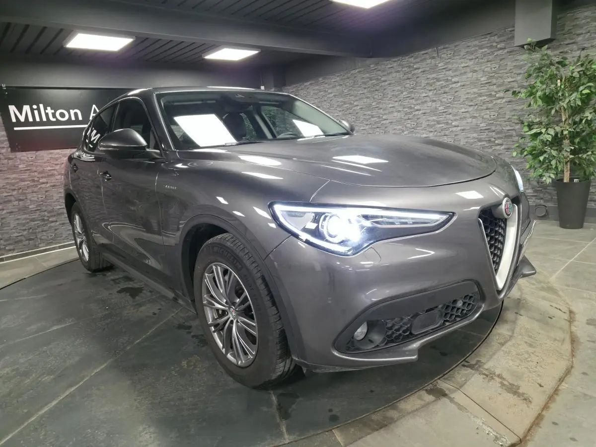 Vue 3/4 avant gauche d'un Alfa Romeo Stelvio gris métallisé avec jantes alliage 18 pouces et phares LED allumés.