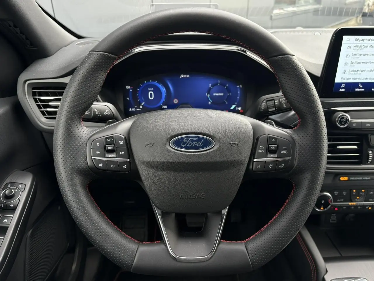 Vue frontale du volant perforé noir avec surpiqûres rouges du Ford Kuga 2023, tableau de bord numérique et écran tactile visibles.