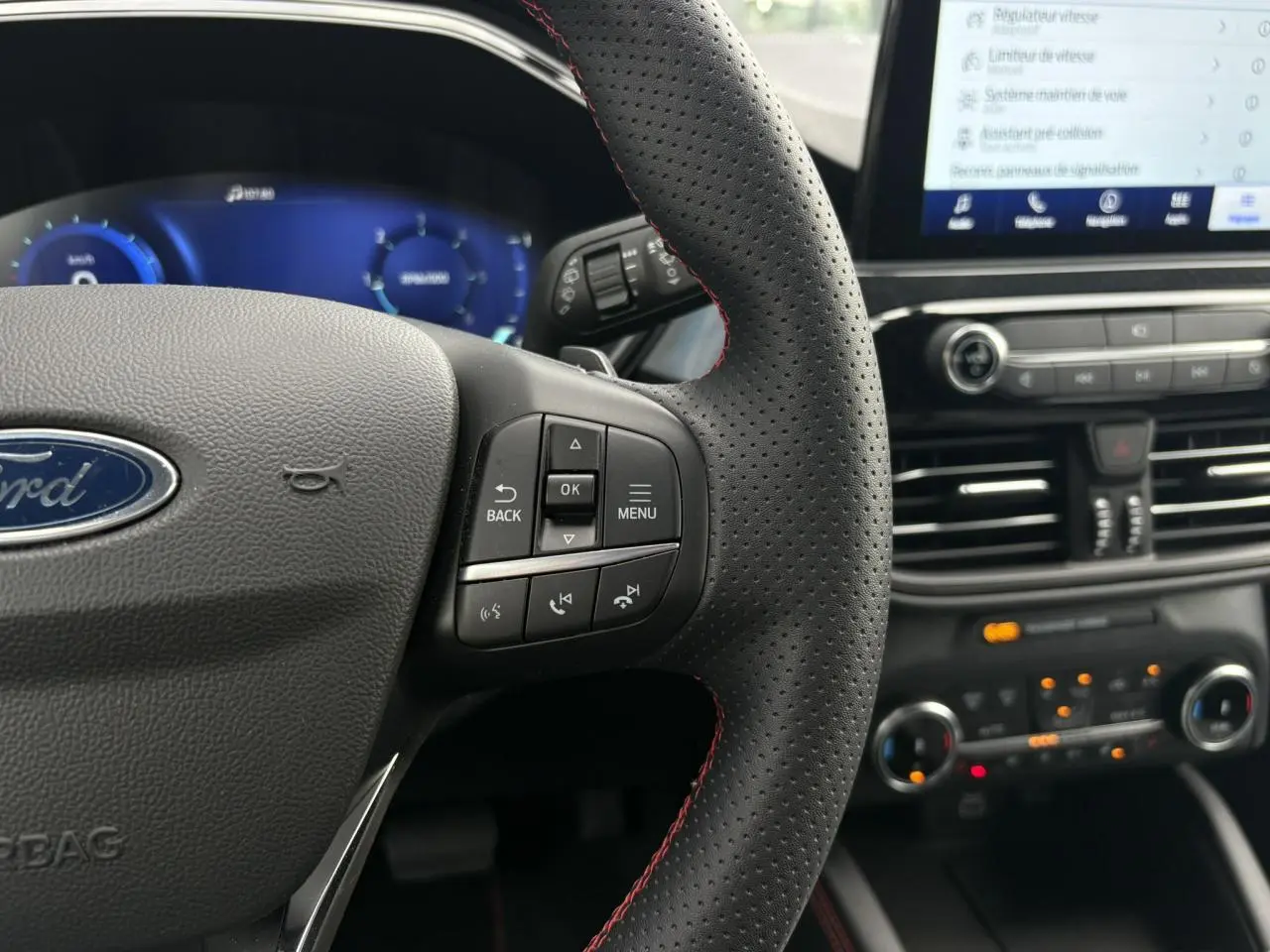 Gros plan sur le volant perforé noir avec surpiqûres rouges du Ford Kuga 2023, tableau de bord numérique et écran tactile visible.