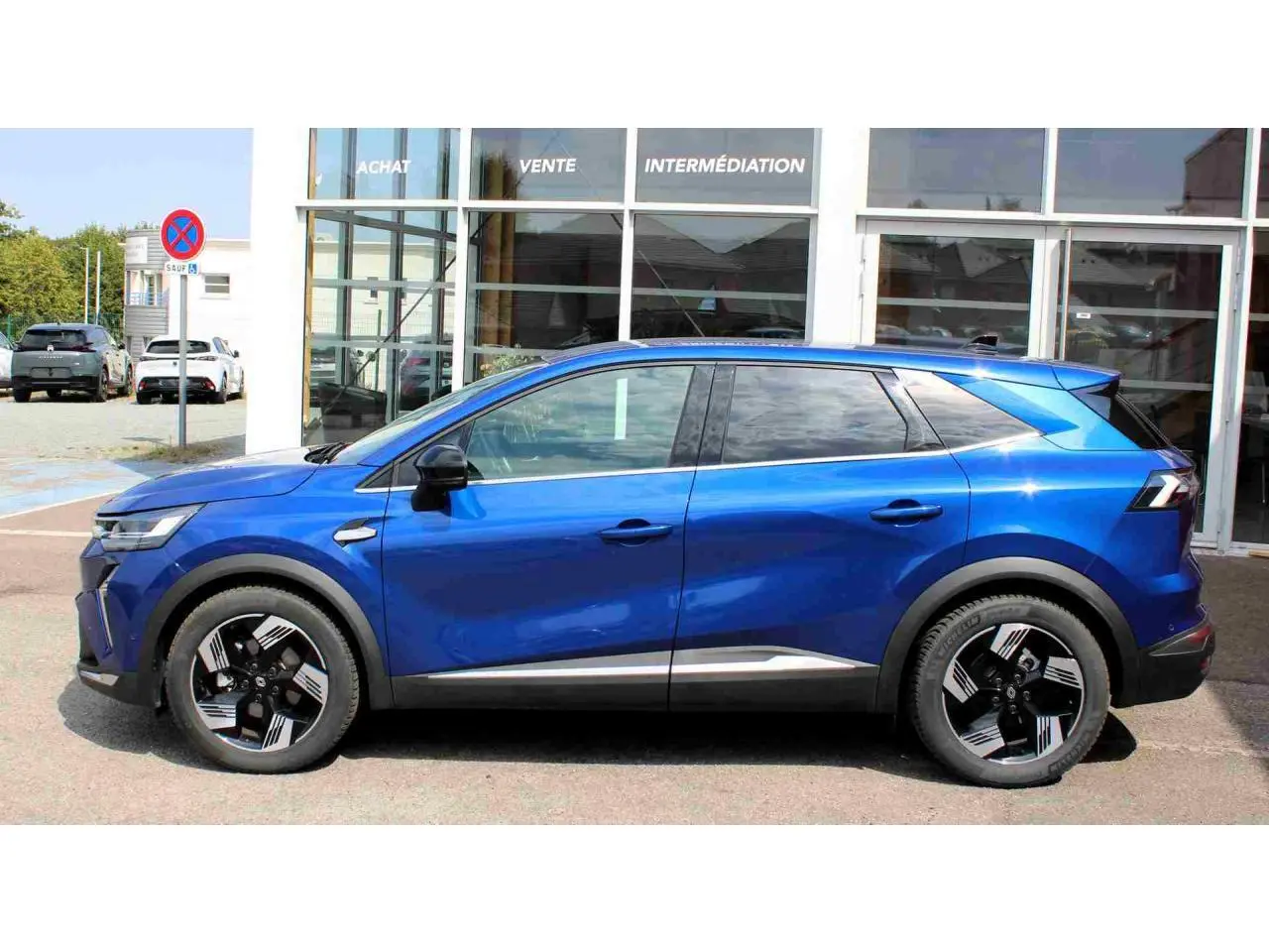 Profil droit d'un Renault SYMBIOZ bleu 2025 avec jantes alliage noires et détails chromés, stationné devant un bâtiment vitré.