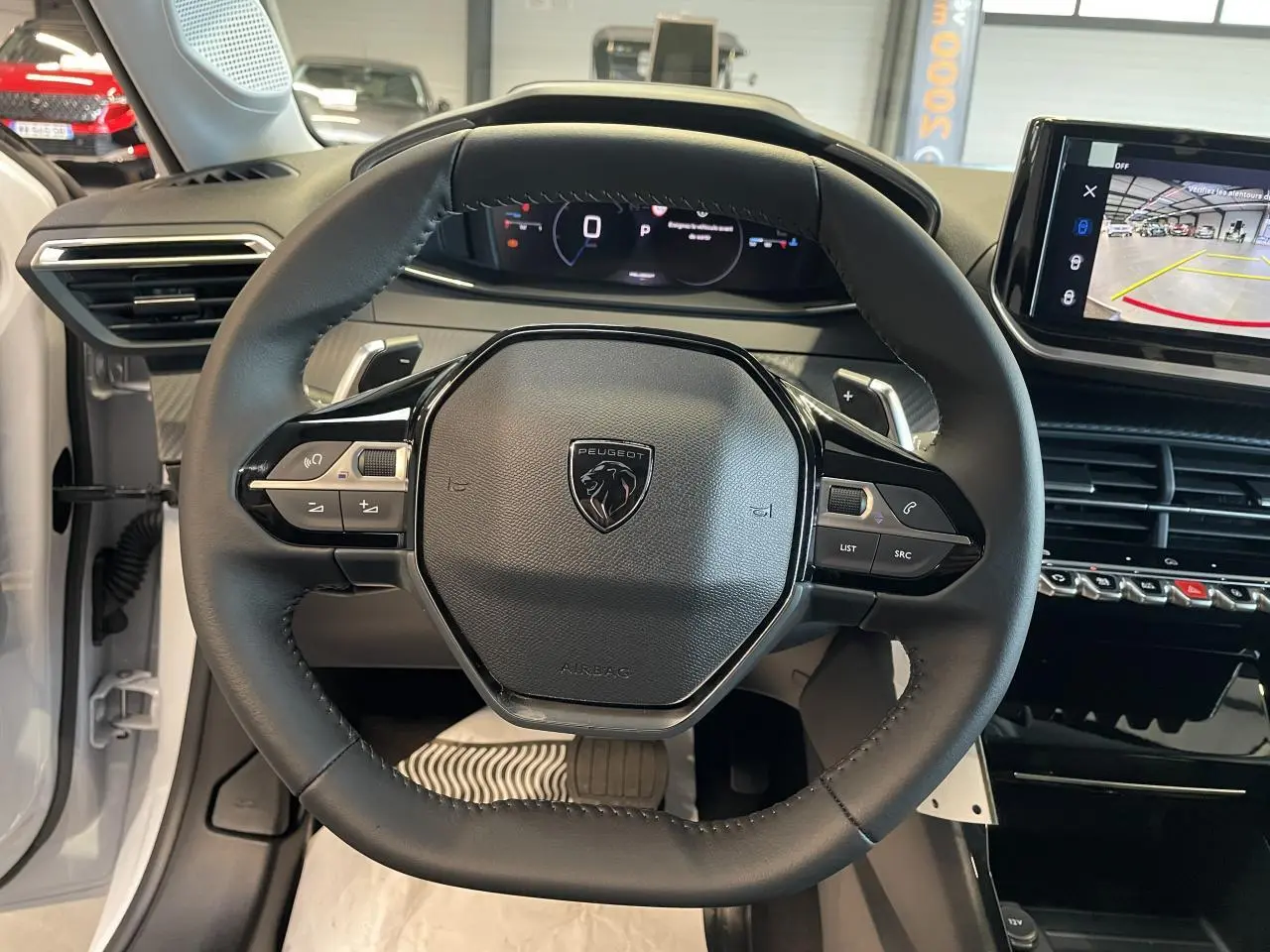 Vue intérieure centrée sur le volant cuir noir avec logo Peugeot, tableau de bord digital et écran tactile en arrière-plan.